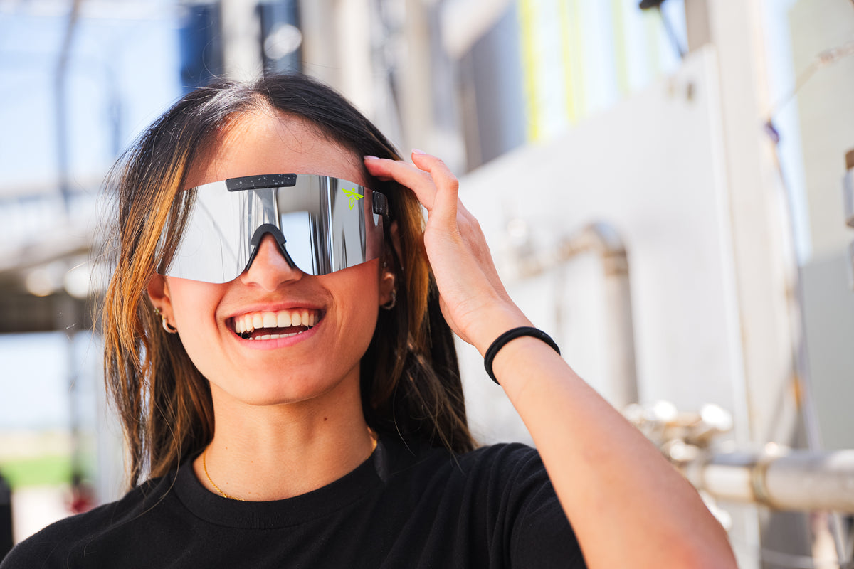 A woman smiling and wearing oversized reflective shield-style sunglasses in an outdoor industrial setting.