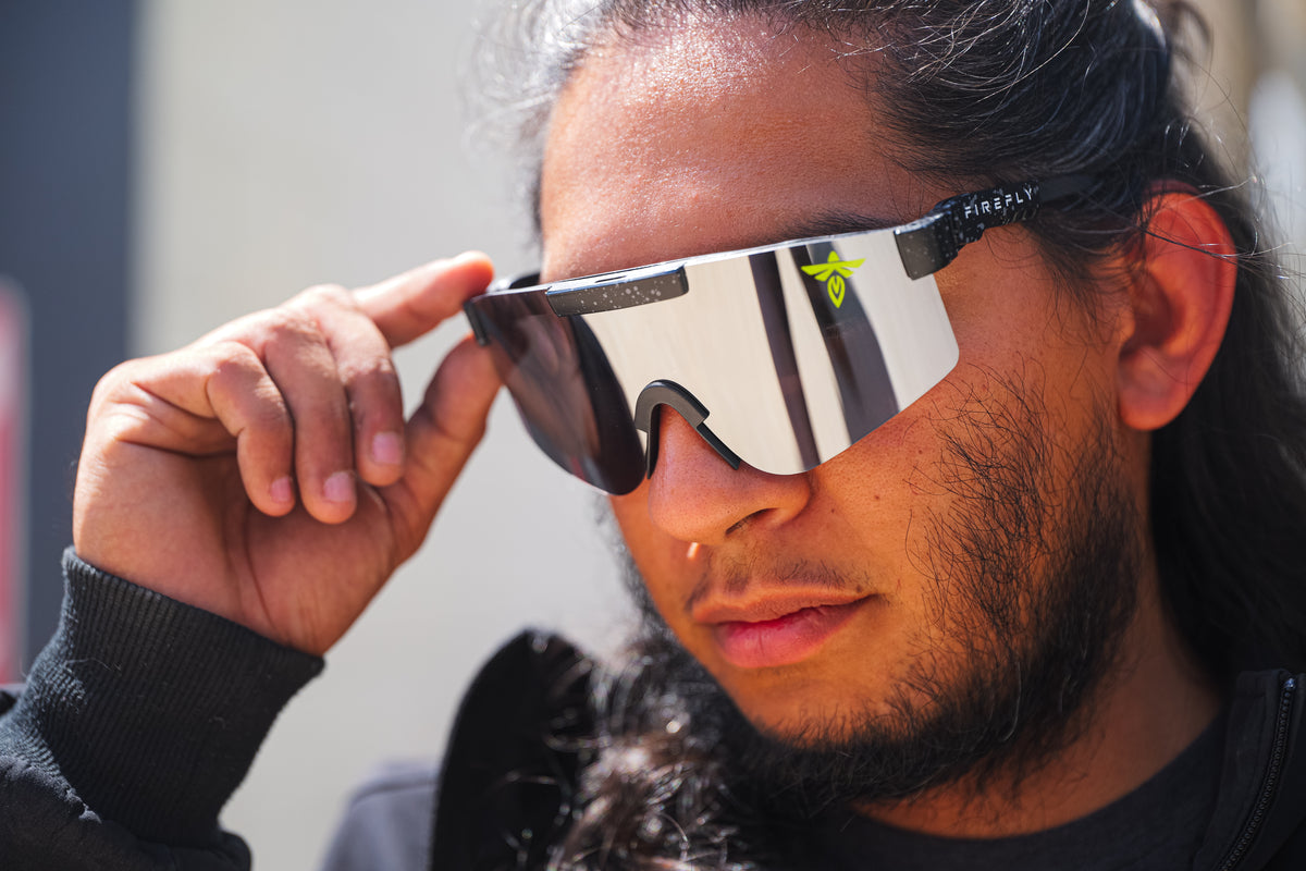 A close-up of a man wearing reflective shield-style sunglasses with a black temple arm and a small neon green logo, holding the side of the glasses with his hand.