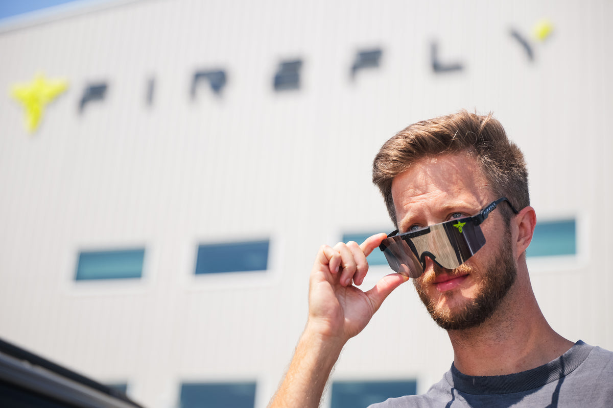 A man outdoors wearing reflective shield-style sunglasses, holding one lens with his hand. A large white building with the word FIREFLY is blurred in the background.