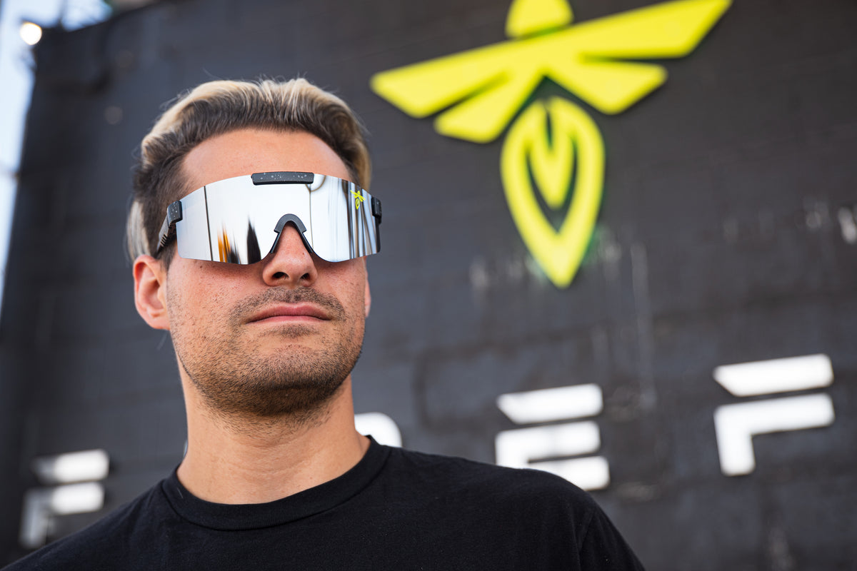A man wearing reflective shield-style sunglasses with a black frame and mirrored lenses, standing outdoors in front of a dark wall with a bright yellow bee logo.