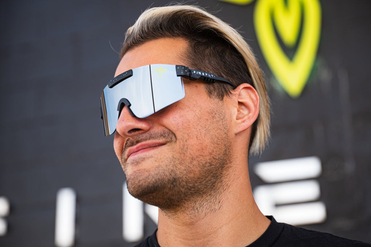 A man wearing oversized shield-style sunglasses with mirrored silver lenses and black arms, shown in a close-up profile against a dark background with a bright yellow logo.