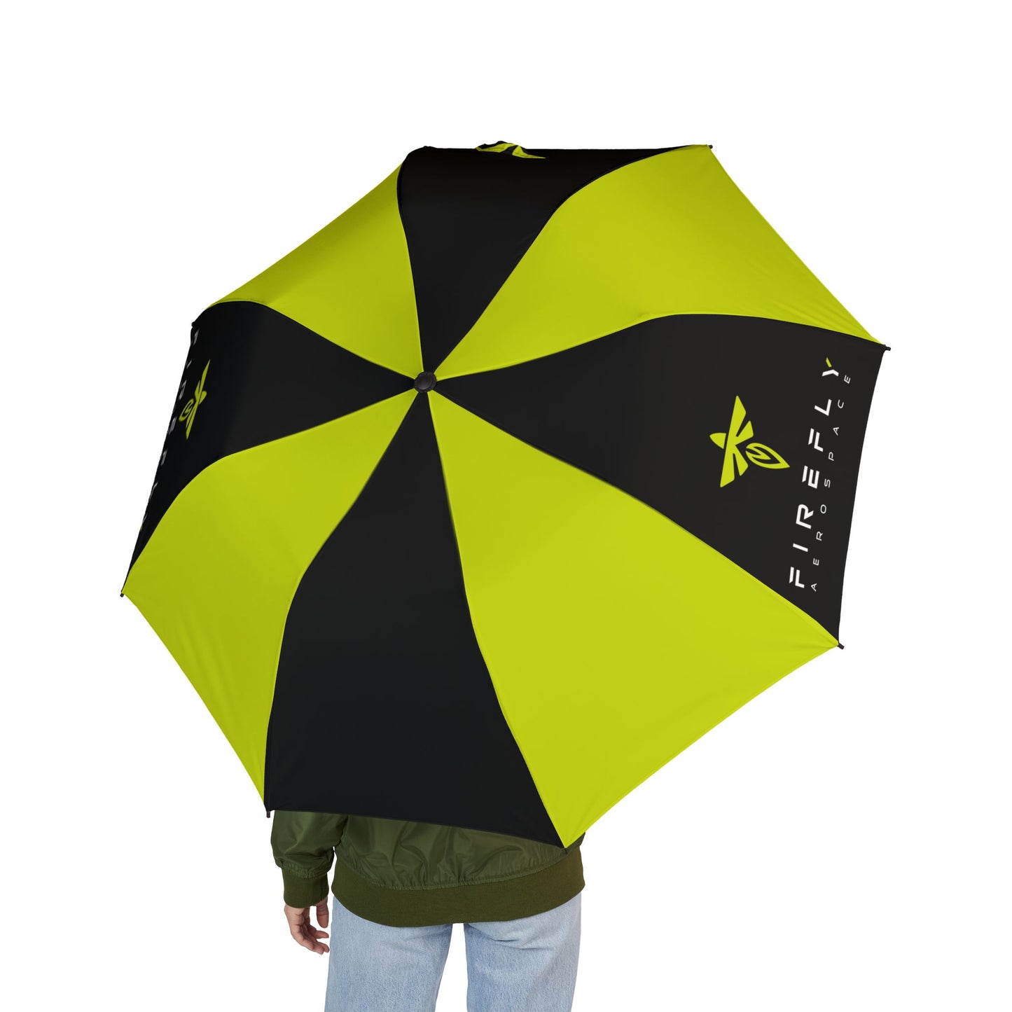A person holding a large umbrella viewed from behind, showing the back of the umbrella with alternating black and bright yellow-green panels, along with printed logos and text. The person is wearing an olive green jacket and light blue jeans against a plain white background.